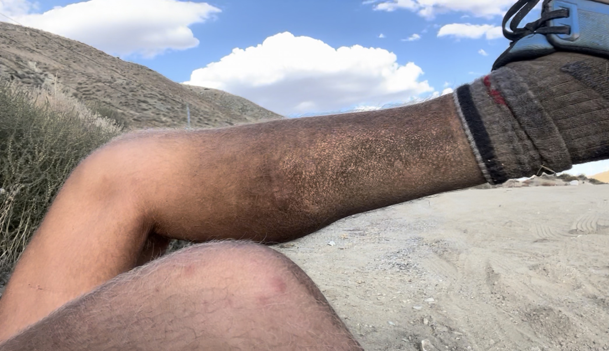A close-up view of two legs resting on a sandy surface, with a mountainous landscape and blue sky in the background.
