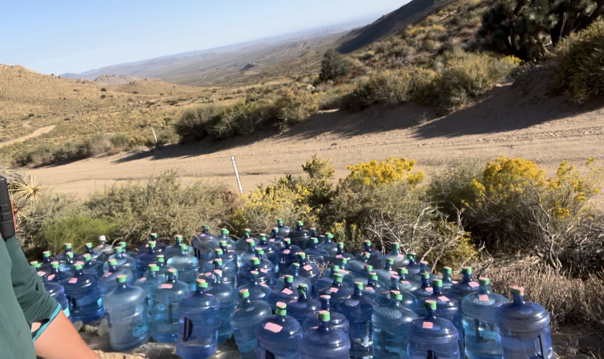 A collection of blue water bottles arranged on the ground, surrounded by desert vegetation and hills in the background.