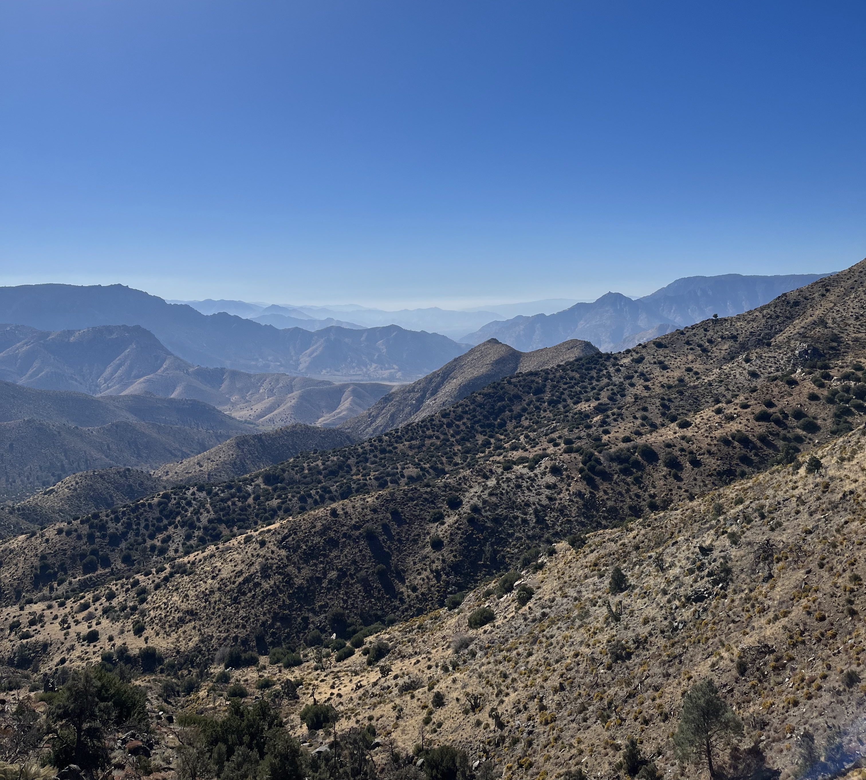 Panoramic view of rolling mountains and hills under a clear blue sky with distant peaks and a landscape featuring sparse vegetation.