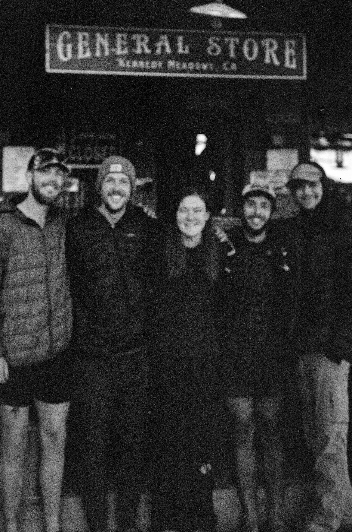 Group of five people standing in front of a general store sign, smiling and posing together.
